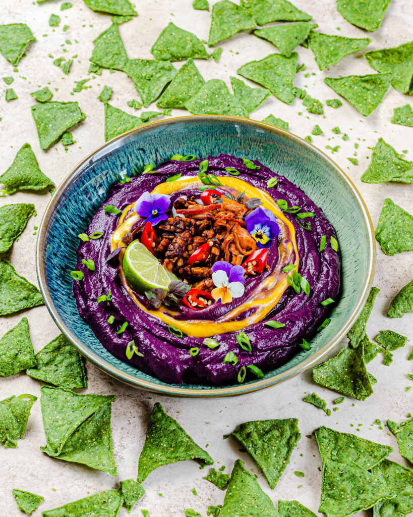 A styled bowl with a purple carrot dip and toppings, surrounded by seaweed chips.