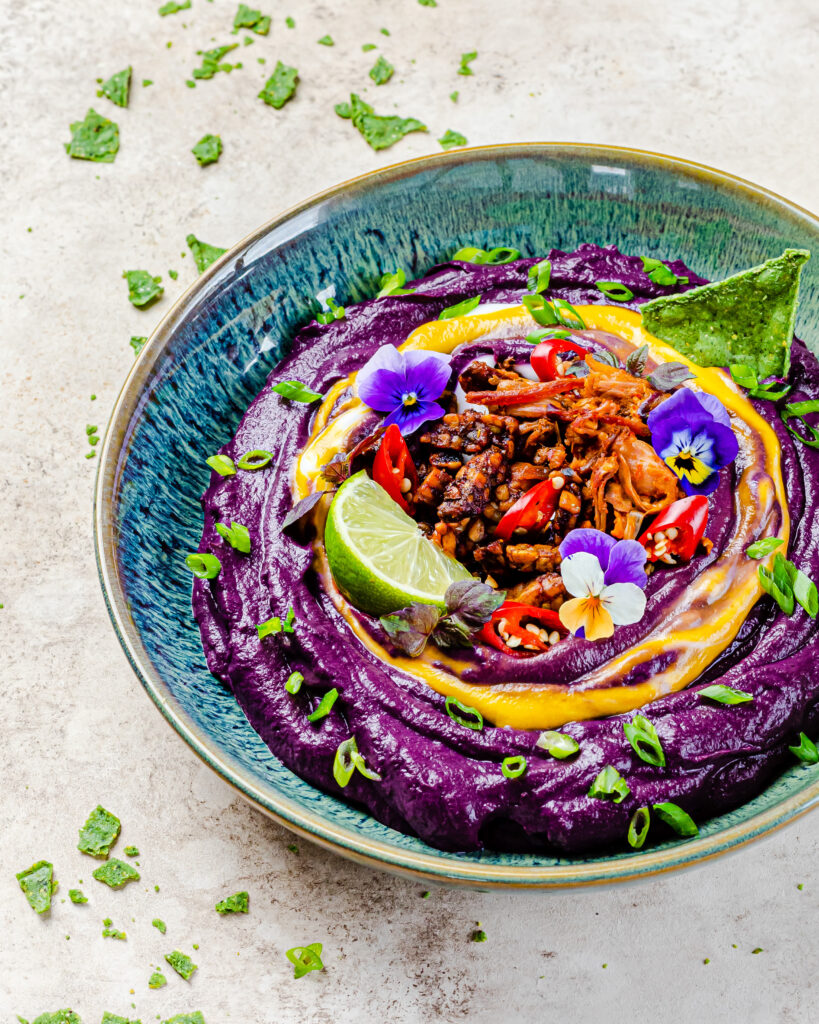 A close up of the purple carrot dip displaying all the toppings. The toppings are sweet and spicy tempeh, bbq jackfruit, scallion, edible flowers and a yellow coconut yoghurt swirl.